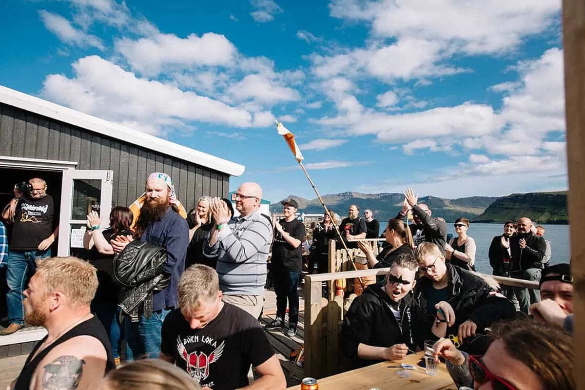 Gestir samankomnir á sólríkri verönd við sjóinn með fjöllin í baksýn.